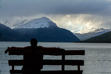 silhueta de uma pessoa sentada em um banco de madeira à beira do lago com montanhas cobertas de gelo ao fundo