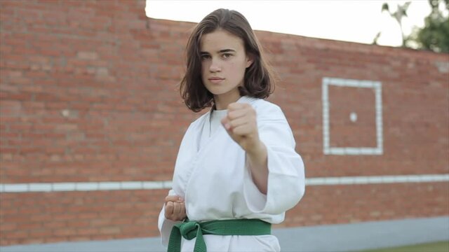 Young woman in a white kimono stands in a basic karate stance on the sports ground and looks at the camera. Front view. Close-up. The camera moves from side to side
