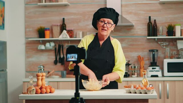 Baker wearing bonete recording podcast of traditional food recipe using flour and cracked eggs. Influencer chef using internet technology communicating, shooting on social media with digital equipment