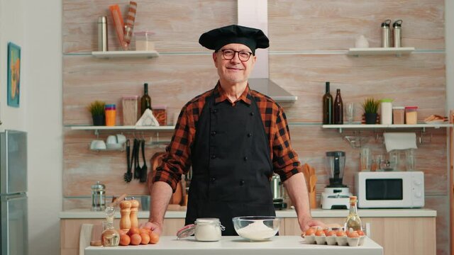 Portrait of chef wearing bonete looking at camera and smiling. Retired elderly baker in kitchen uniform preparing pastry ingredients on wooden table ready to cook homemade tasty bread, cakes and pasta
