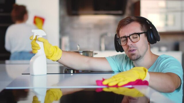 Cheerful man in headphones wiping cooking top with rag and wife cleaning furniture on background