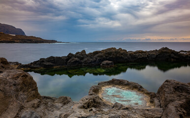 Dark clouds before a sunset on the rocky north coast of Tenerife. A natural pool is separated from the sea by rocks. You can see some red sunset on the horizon.