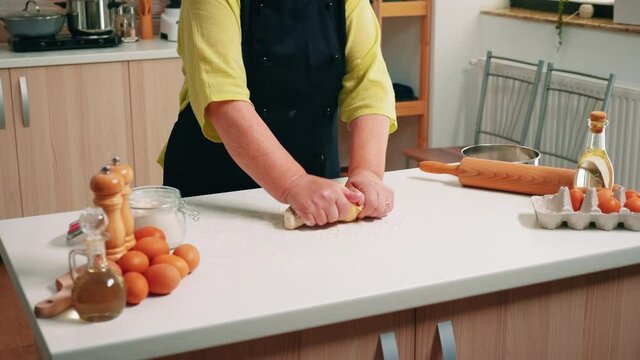 Senior woman chef kneads the dough on table in modern kitchen. Retired elderly baker with bonete mixing ingredients with sifted wheat flour kneading for baking traditional cake and bread.