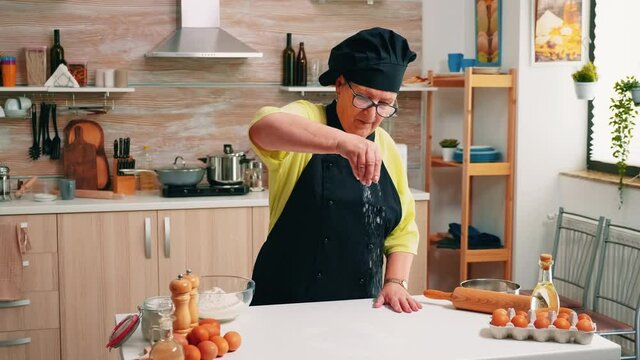Woman with bonete sieving flour on wooden table ready for cooking. Retired senior baker with apron, kitchen uniform sprinkling, sifting, spreading rew ingredients by hand baking homemade bread.