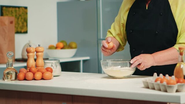 Elderly baker woman cracking eggs on glass bowl for tasty food recipe in home kitchen. Retired elderly chef with bonete mixing by hand, kneading pastry ingredients baking homemade cake.