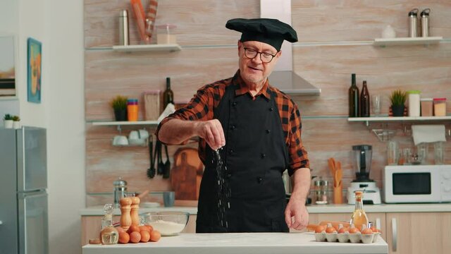 Senior baker in home kitchen putting wheat flour over wooden table. Retired elderly chef with bonete and apron sprinkling, sieving sifting raw ingredients by hand baking homemade pizza, bread.