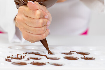 Close-up chocolatier pours hot dark chocolate into confectionery mold using pastry bag, filling out...