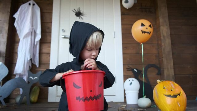 Child halloween portrait. Cute little kid in wizard costume sitting on porch of house, holding basket and looking at how much candy he collected while trick-or-treating. Handmade diy holiday decor.