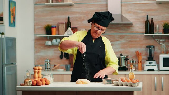 Skilled old woman reparing dough for baking in home modern kitchen. Retired senior chef with bonete and uniform sprinkling, sieving sifting wheat flour with hand baking homemade pizza and bread.