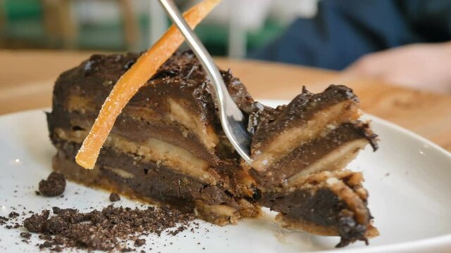 Vegetarian cafe visitor inputs silver fork into vegan chocolate cake slice sitting at brown wooden table slow motion closeup