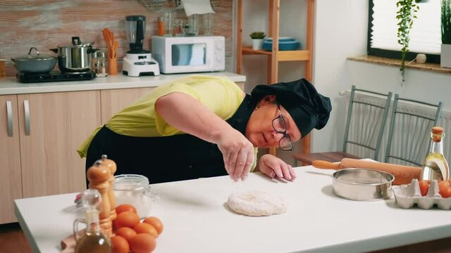 Floured pizza dough on wooden table ready to be cooked. Elderly senior baker with bonete and uniform sprinkling, sifting, spreading rew ingredients on dough by hand, baking homemade cakes and bread.