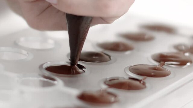cropped view of chocolatier hand with pastry bag pouring melted dark chocolate into candy mold preparing luxurious handmade confectionery slow mo. World Chocolate Day