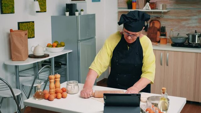 Old woman with bonete participating at culinary master class on laptop. Retired chef following baker advice on tablet, learning cooking tutorial on social media, using rolling pin forming the dough.