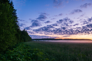 A big blue sky with a beautiful orange sunset. Green trees in the foreground, the beginning of the forest, the white mist in the field.