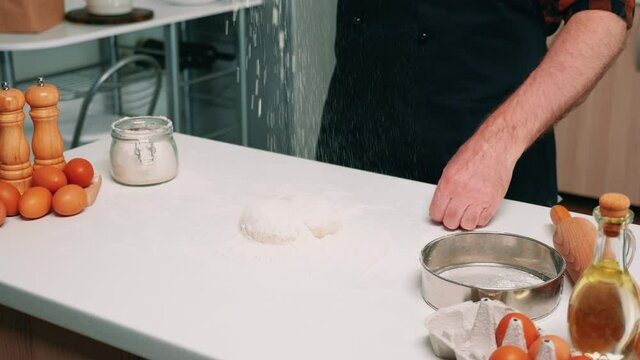Hands of chef man sifting flour on dough for baking. Elderly senior baker with bonete and uniform sprinkling, sieving, spreading rew ingredients by hand cooking homemade pizza and bread.