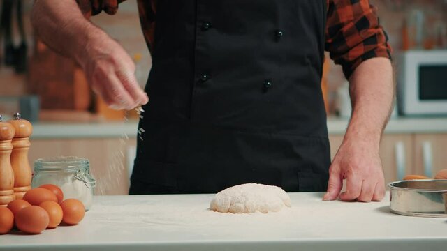 Senior bakery man sifting wheat flour on dough for kneading. Retired elderly chef with bonete and uniform sprinkling, sieving spreading rew ingredients with hand baking homemade pizza and bread.