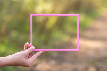Hand holding a empty, wooden, pink frame on nature background. Care, safety, memory or painting concept. The concept of creativity and inspiration. Space for text. Mock up