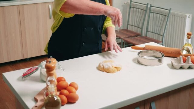Retired woman chef cooking on wooden table in home kitchen. Elderly senior baker with bonete and uniform sprinkling, sifting, spreading rew ingredients on dough, baking homemade pizza and bread.