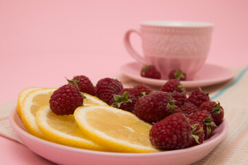 lemon wedges and raspberries in a pink plate, a cup of tea on a pink background. Flat lay of tropical summer. Food concept. Disease prevention.