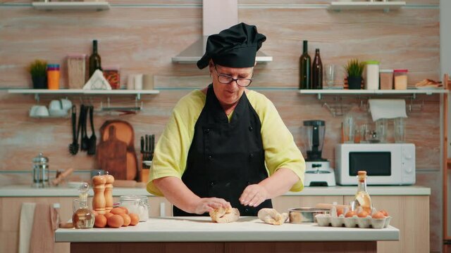 Senior woman rolling out the dough preparing homemade bread. Happy elderly chef with bonete preparing raw ingredients to baking traditional pizza sprinkling, sieving flour on table in the kitchen.