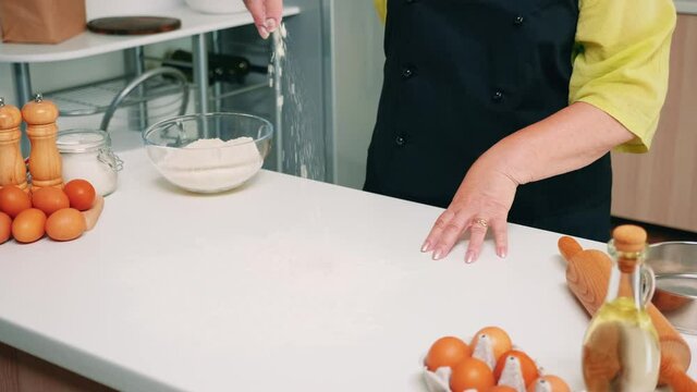 Making dough by woman hands at home in kitchen using wheat flour. Retired senior baker with bonete and uniform sprinkling, sifting, spreading rew ingredients baking homemade pizza and bread.