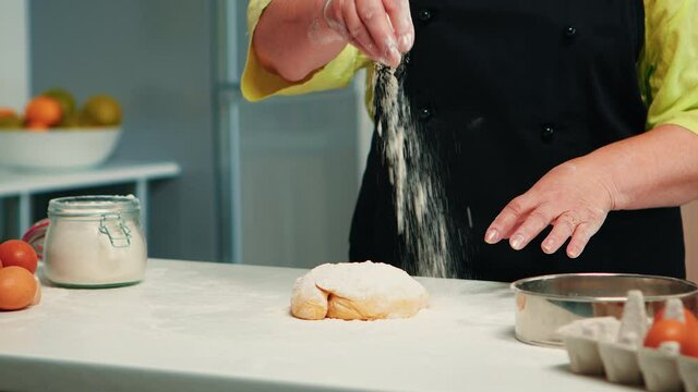 Grandmother preparing homemade donuts wearing kitchen apron. Retired senior chef with bonete and uniform sprinkling, sieving sifting wheat flour with hand baking homemade pizza and bread.