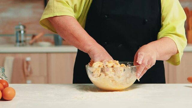 Woman preparing tasty food using wheat flour and cracked fresh eggs in home. Close up of retired elderly chef with bonete kneading in glass bowl pastry ingredients baking homemade cake and bread.