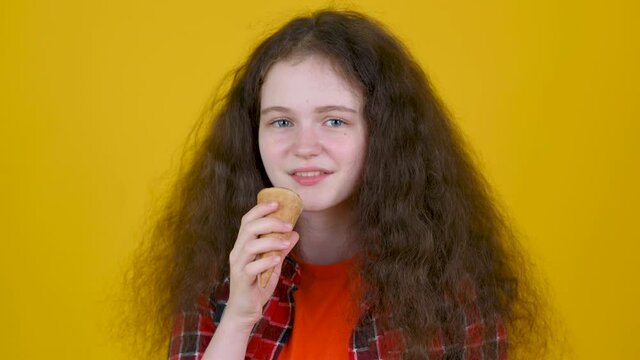 Close up portrait of young girl with curly hair in shirt eating ice cream isolated on yellow studio background
