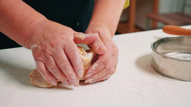 Woman hands forming loaf of bread near kitchenware sprinkled with flour table. Retired elderly baker with bonete mixing ingredients with sifted wheat flour kneading for baking traditional cake, bread
