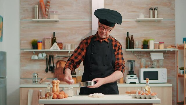 Baker with bonete and apron using metallic sieve preparing bakery products at home. Happy elderly chef with kitchen uniform mixing, sprinkling, sieving raw ingredients to baking traditional bread