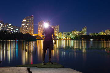 Fototapeta na wymiar A guy stands on the banks of the Dnieper river against the background of a bright night embankment