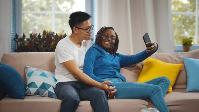 Young diverse joyful couple hugging and smiling while taking selfie photo in living room