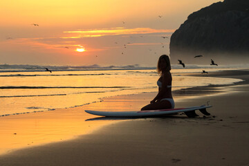 Woman sitting on the sunset beach with surfboard