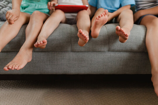 Anonymous children's bare feet dangling from sofa while playing on an electronic device indoors in the summer