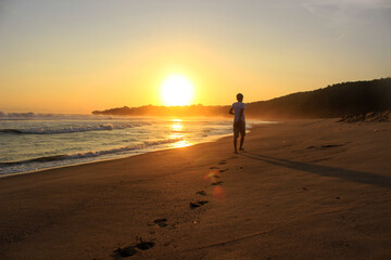 Naklejka premium A man walks toward the setting sun, leaving footprints in the sand. Far look from behind. 