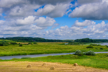 Fototapeta na wymiar Stunning landscape with river, field, haystacks and gorgeous clouds in the sky