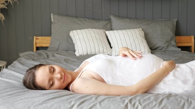 Happy pregnant woman in white dress lies on bed stroking tummy at home close-up