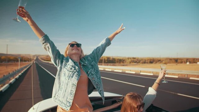 Two young beautiful women on a cabriolet with a cabriolet top drink champagne and have fun during their trip. Friendship, vacation, youth holidays lifestyle concept