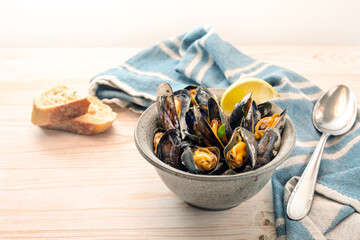 Bowl with mussels, lemon and herbs, a blue towel, spoon and bread on a light wooden table, copy space