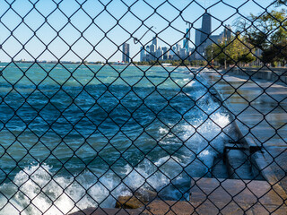 view through fence of waves spraying up on lakefront of Lake Michigan with view of downtown Chicago