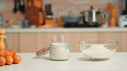 Glass bowl of wheat flour and fresh eggs on table in empty kitchen. Modern dining room equipped with utensils ready for cooking with pastry ingredients for homemade cakes and bread