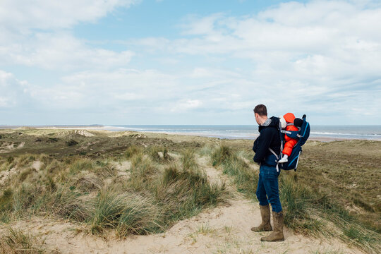 Young father walking on the coast with his daughter in a baby carrier backpack. Norfolk, UK.