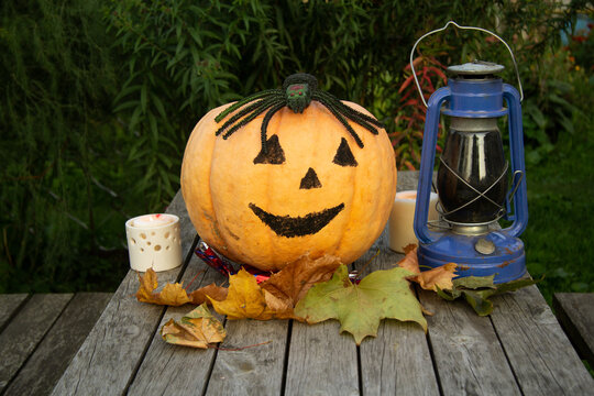 A large pumpkin with painted eyes and mouth lies on a wooden table. Nearby are candles and a kerasin lamp. On the pumpkin lies a large toy rubber spider