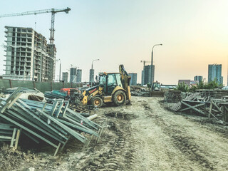 construction site of a new district. yellow construction equipment and building materials on site. against the background of tall houses. construction cranes are building a neighborhood