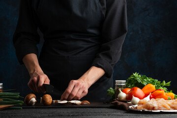 The chef in black uniform cuts with knife mushrooms on the black chopping board at the professional kitchen on dark blue background. Backstage of preparing restaurant dish. Food concept.
