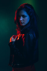 Night portrait. Neon light. Female mystery. Anxiety pressure. Pensive insecure brunette woman in leather biker jacket standing in red blue glow isolated on dark teal green background.