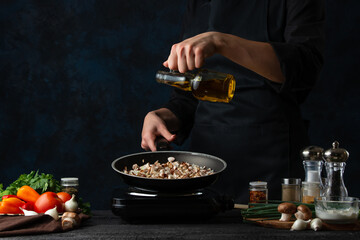 The chef in black apron pours oil into the pan with frying meat on dark blue background. Professional kitchen table with spices, vegetables, electric stove and other ingredients for cooking.
