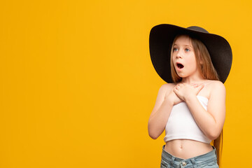 Impressed child. Holiday surprise. Wow reaction. Big chance. Portrait of astonished young girl in black elegant summer hat looking at copy space isolated on orange promotional background.