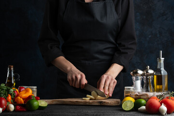 The chef in black uniform cuts with knife potato on the wooden chopping board at the professional restaurant kitchen. Dark blue background. Backstage of preparing dish with potato. Eco-friendly food.