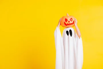Tuinposter Funny Halloween Kid Concept, little cute child with white dressed costume halloween ghost scary he holding orange pumpkin ghost on hand, studio shot yellow on white background © sorapop